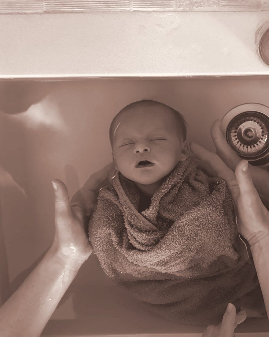 Bébé emmailloté qui bâille, la tête soutenue par des mains dans l'eau du bain